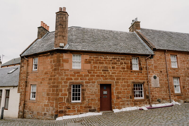 A red sandstone house with tall chimneys stands on a cobbled street corner, a plaque fixed to its wall beside a window.