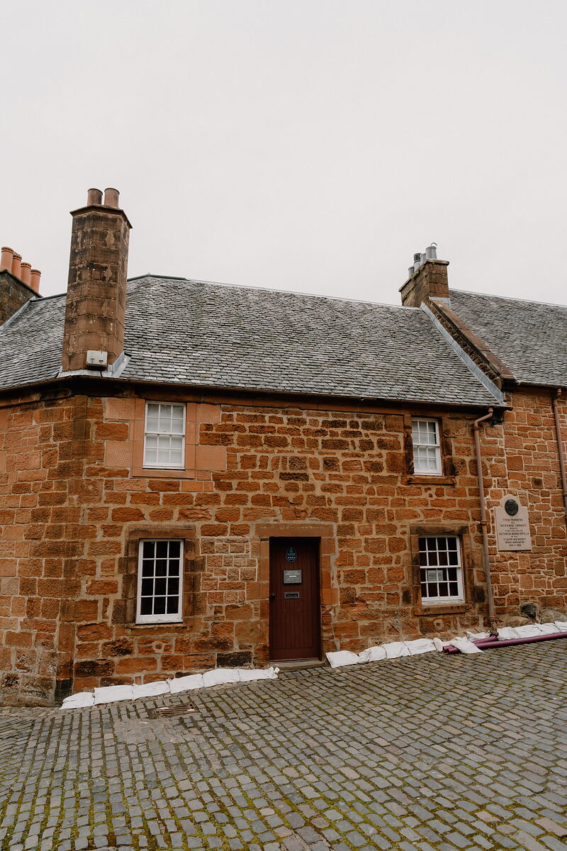 A red sandstone cottage with tall chimneys stands on a cobbled street, sandbags lined along its base and a small plaque beside the door.