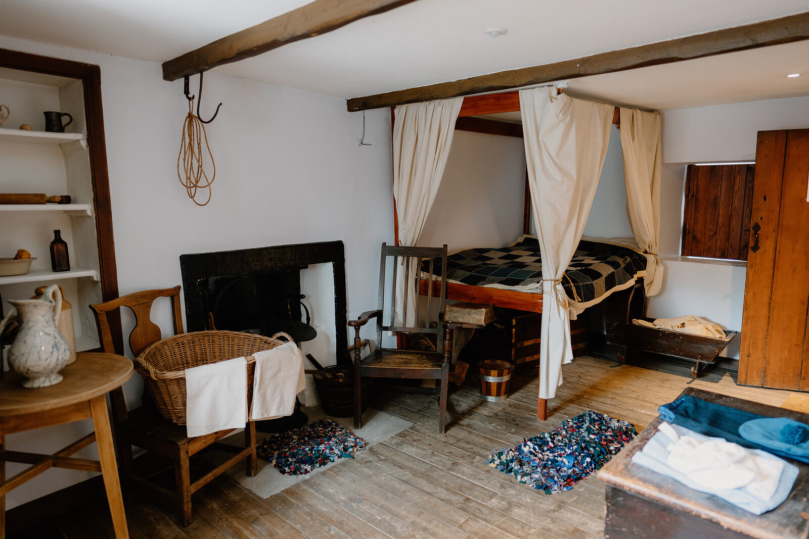 A box bed with pale curtains stands in the corner of a whitewashed cottage room, a fireplace, wicker chair and patterned rugs completing the furnishings.