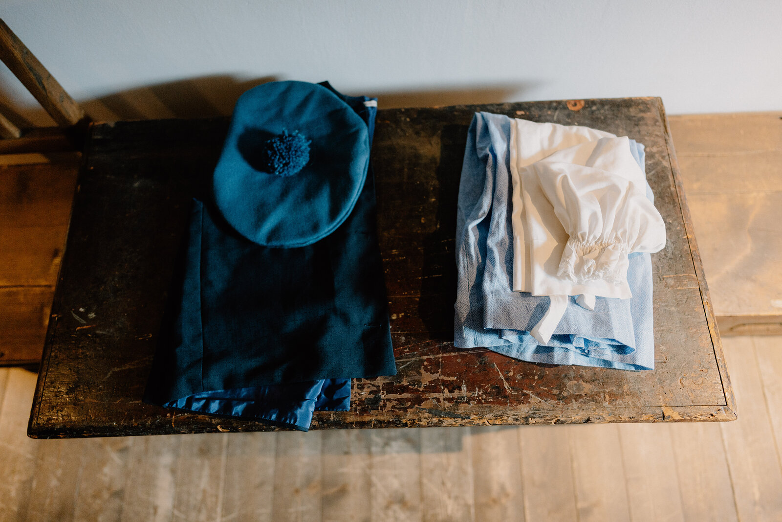 A dark blue bonnet and cape lie folded beside pale striped clothing on an old wooden table.