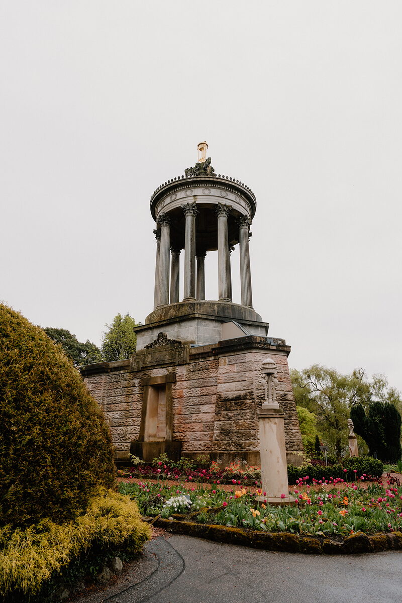 A domed stone monument with tall columns rises above a bed of tulips and a low box hedge, a stone plinth standing to one side.