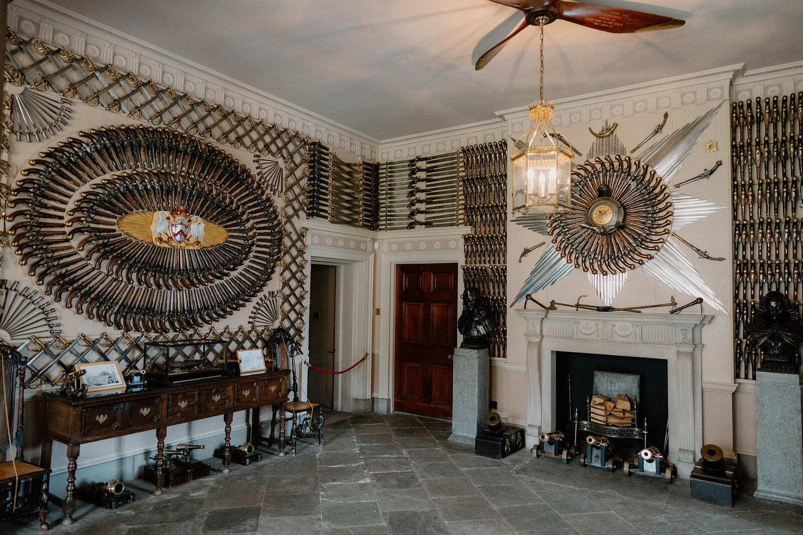 An oval and star pattern of antlers and weapons covers the walls of a grand room, a stone fireplace and dark wooden furniture standing below.