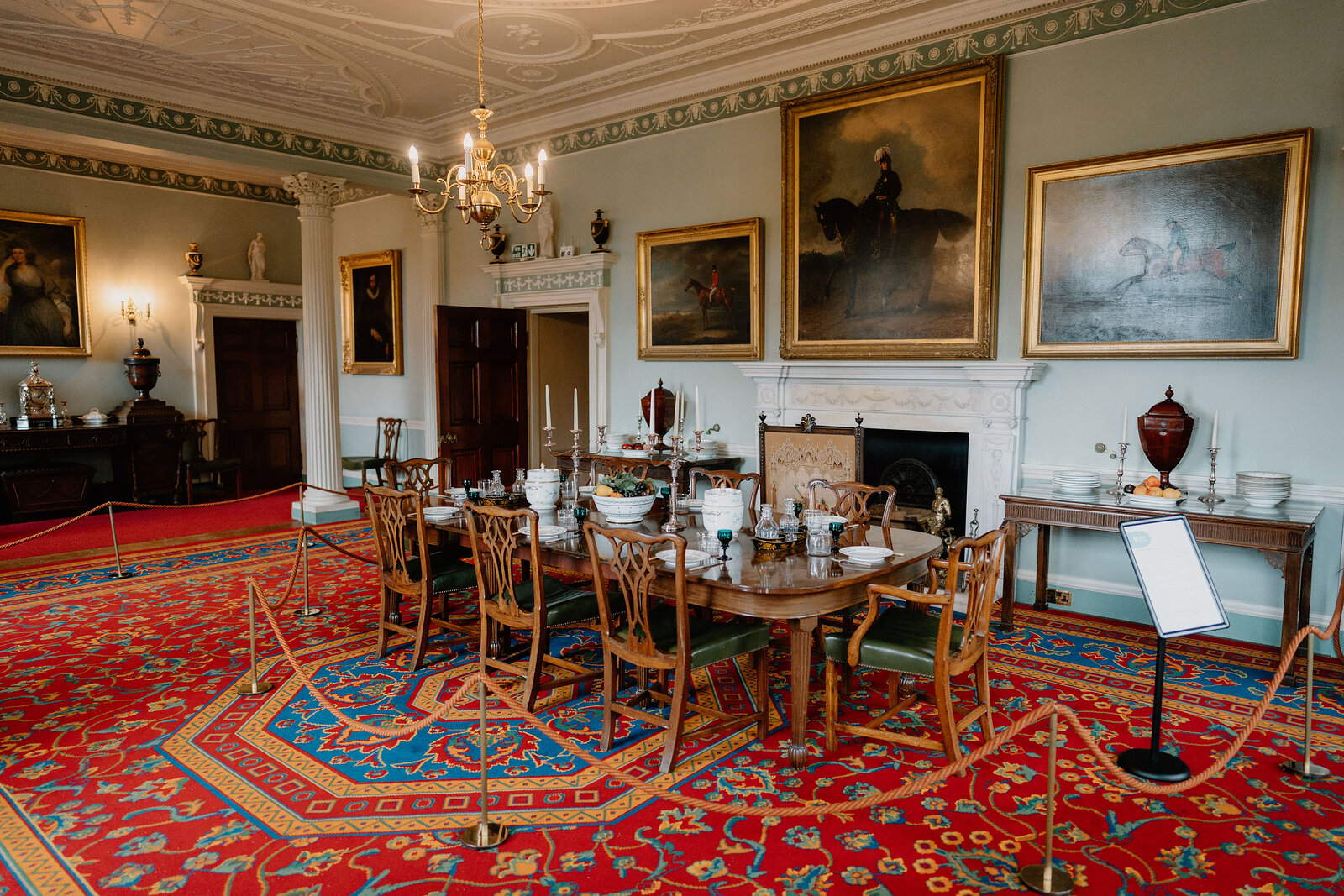 A grand dining room with a red patterned carpet, a chandelier hanging from an ornate ceiling, oil paintings above a marble fireplace, and a long table laid with china and decanters on a sideboard behind a rope barrier.