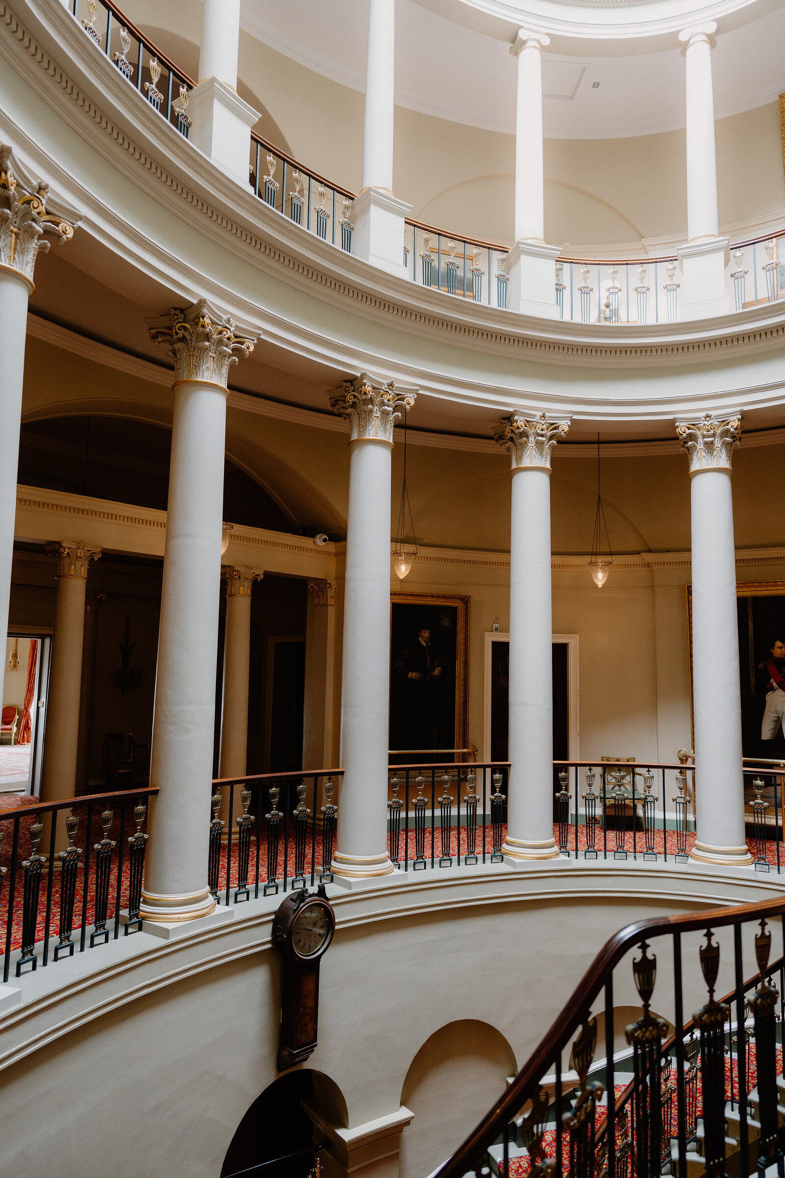 A ring of Corinthian columns rises through a domed rotunda past a galleried landing hung with portraits, a wall clock mounted on the curved staircase below.
