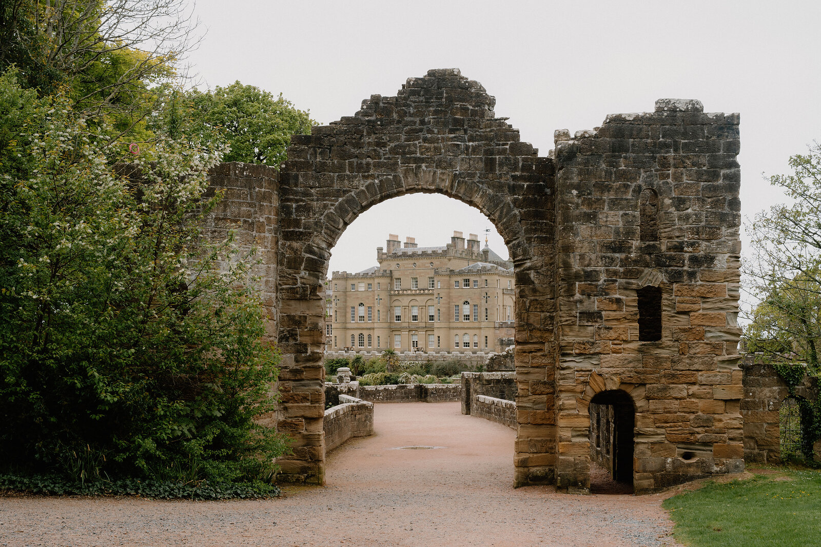 A ruined stone archway frames a distant view of a grand stone mansion at the end of a gravel drive, trees and lawn either side.
