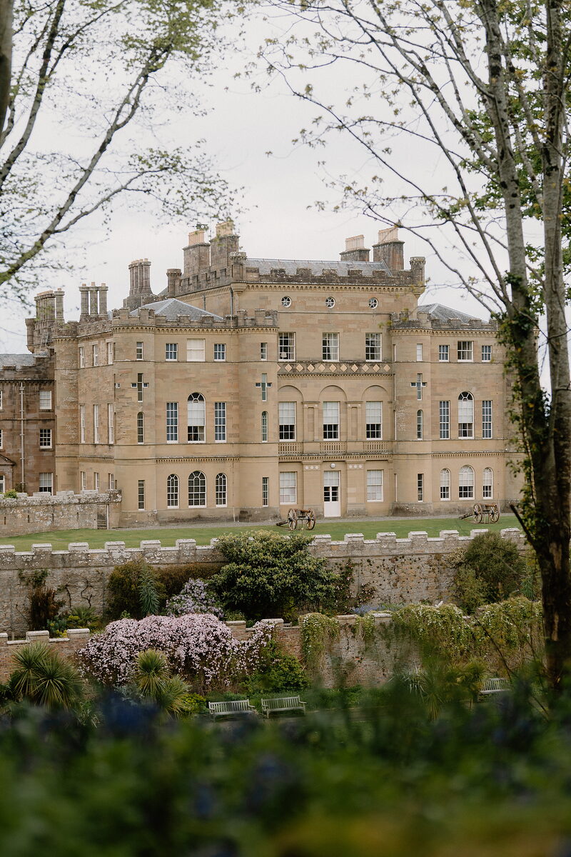 A castellated stone mansion with rows of chimneys stands behind a walled lawn where two old cannons point outward, trees and blossom framing the view.