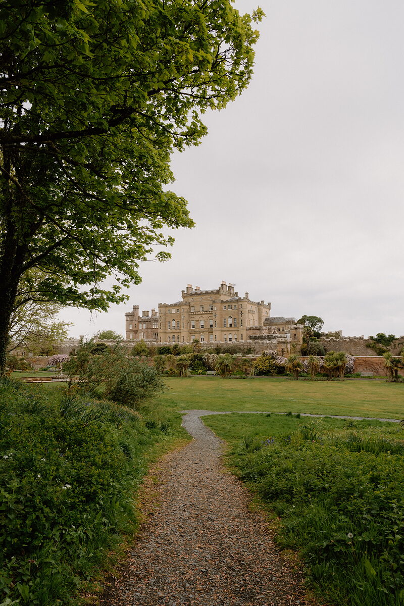 A castellated stone mansion with rows of chimneys stands beyond a lawn, a gravel path curving through shrubs and grass toward it.