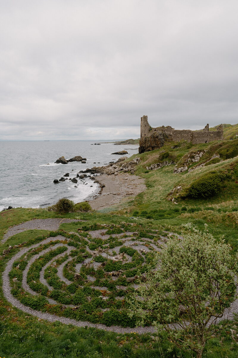 A spiral turf maze sits on a grassy clifftop above a pebble beach, the ruined walls of a coastal castle standing further along the shore.