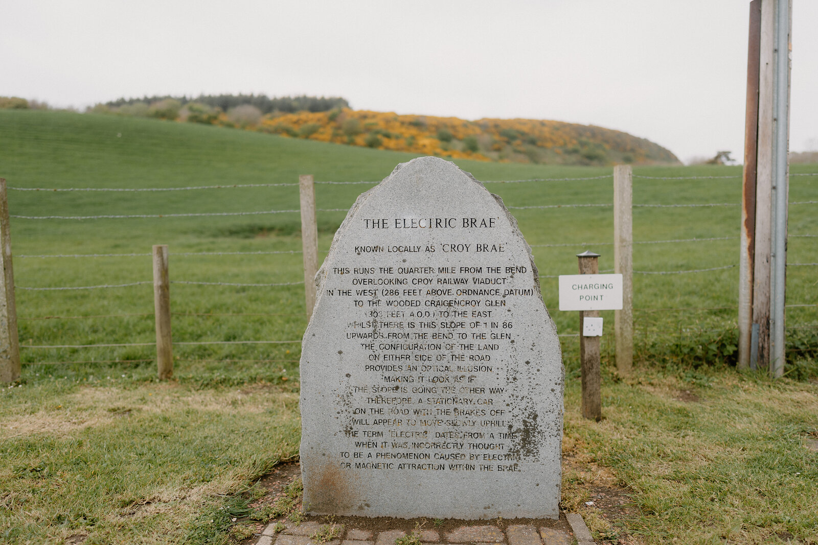 A carved stone monument reading "The Electric Brae, known locally as Croy Brae" explains the road's optical illusion, an electric vehicle charging point standing beside it.