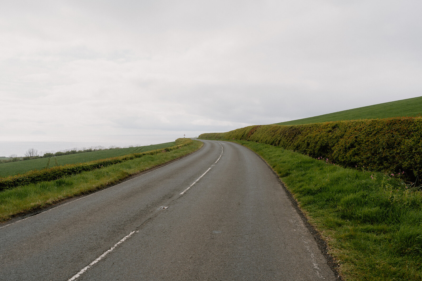 A country road curves between hedgerows along a coastal hillside under an overcast sky.