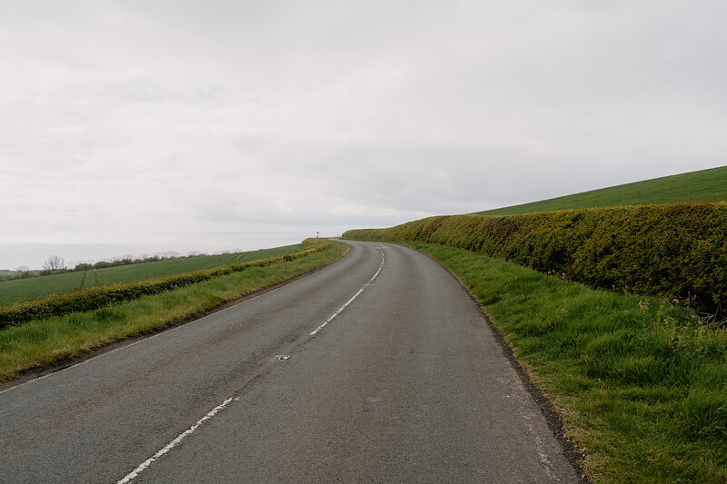 A country road curves between hedgerows along a coastal hillside under an overcast sky.