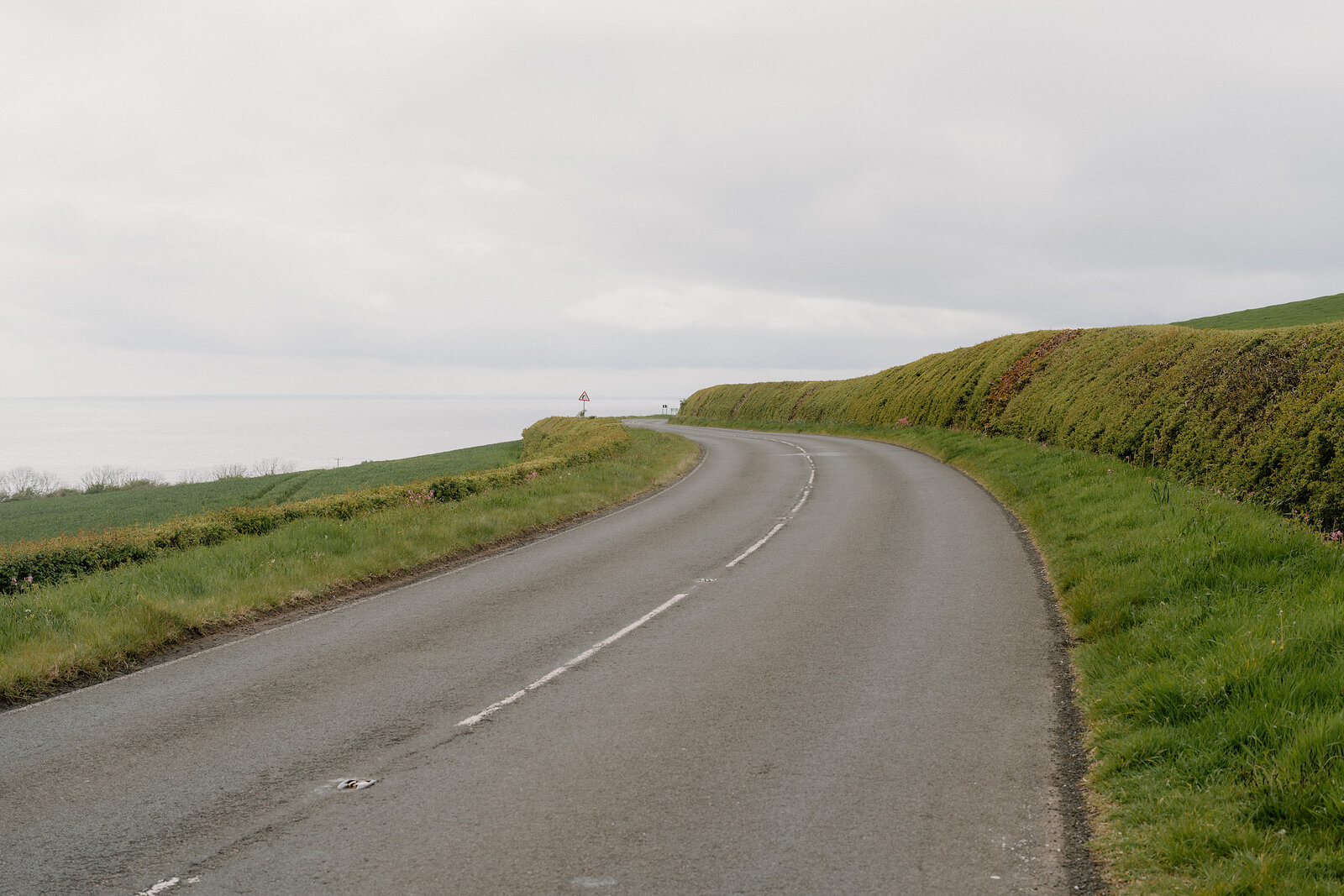 A country road curves between hedgerows along a coastal hillside under an overcast sky.