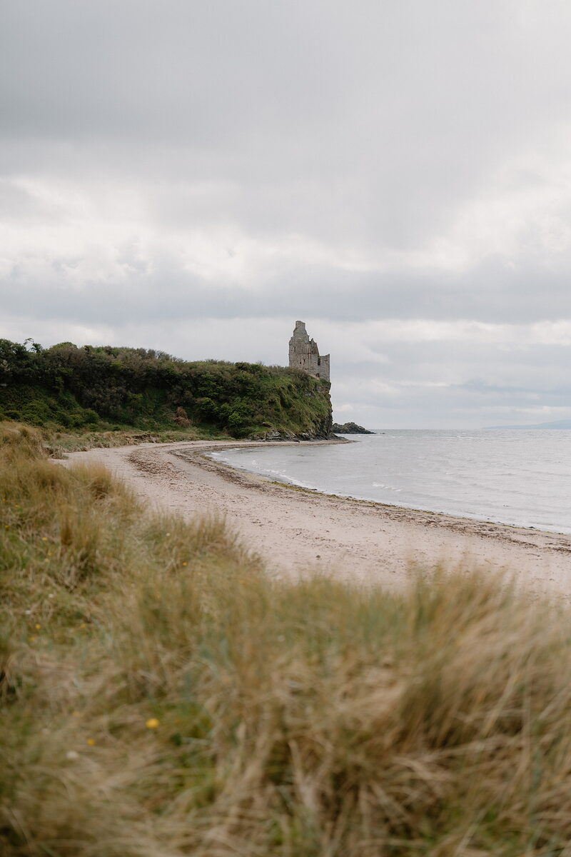 The ruined tower of a coastal castle stands on a grassy clifftop above a curving sandy beach.