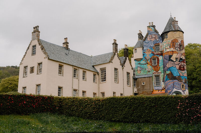 A large mural depicting robed figures covers the round turret of a whitewashed mansion, a clipped hedge bordering the lawn in front.