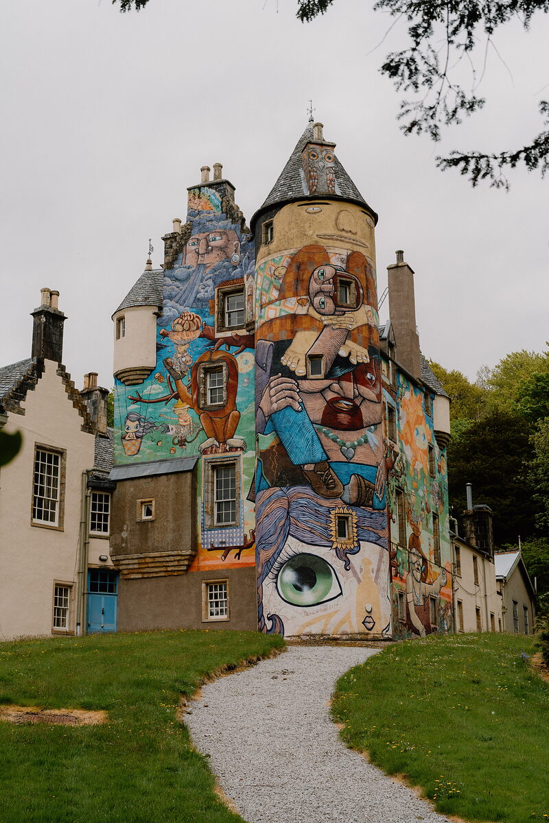 A large mural depicting robed figures and a giant eye covers the turret and gables of a whitewashed mansion, a gravel path leading toward it across a lawn.