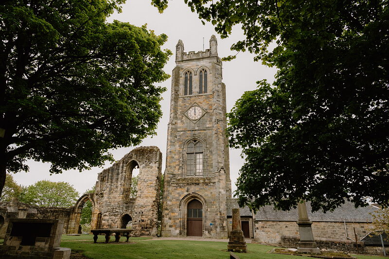 The ruined tower of a Gothic abbey with a clock face rises above trees, benches and gravestones standing in the churchyard below.