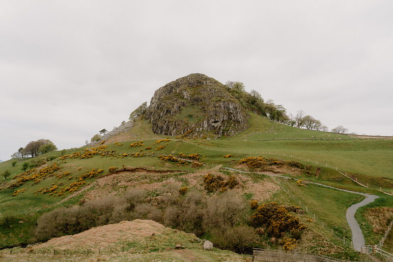 A grass-covered conical hill dotted with gorse rises above a rock outcrop, a path curving around its base.