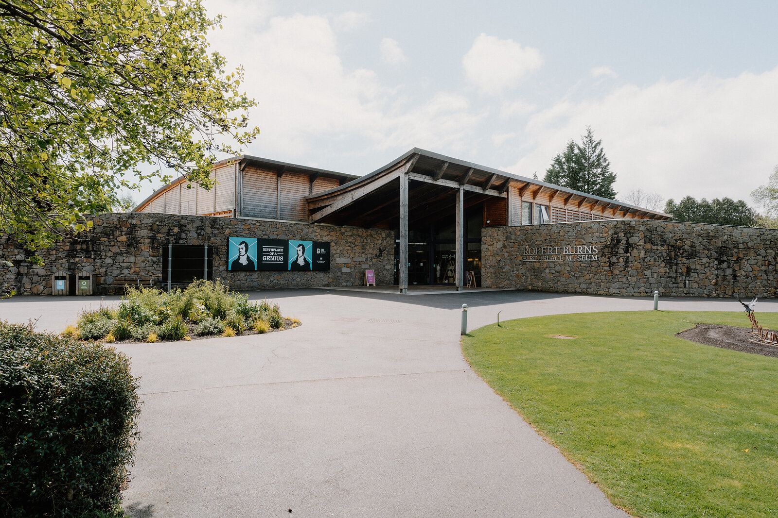 A timber-clad museum building with an angular roofline stands beyond a stone wall, illuminated banner screens and a gravel forecourt in front.