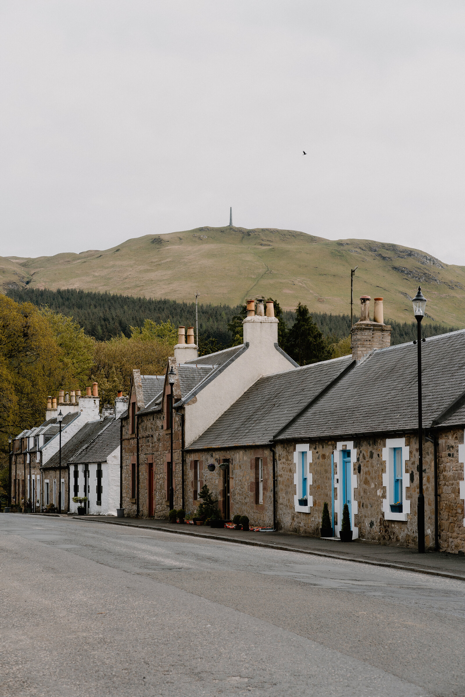 A row of stone cottages with tall chimney pots lines a street, an obelisk memorial standing on the hill behind.
