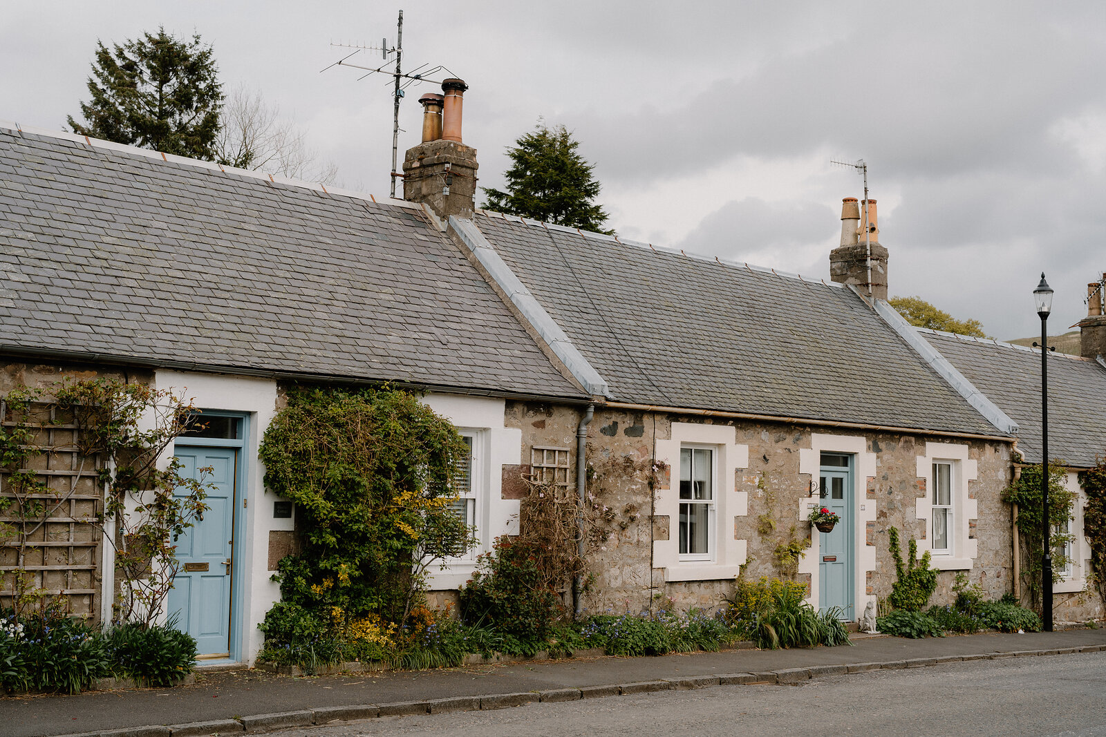 A row of stone cottages with pale blue doors and TV aerials lines a street, climbing plants softening the frontages.