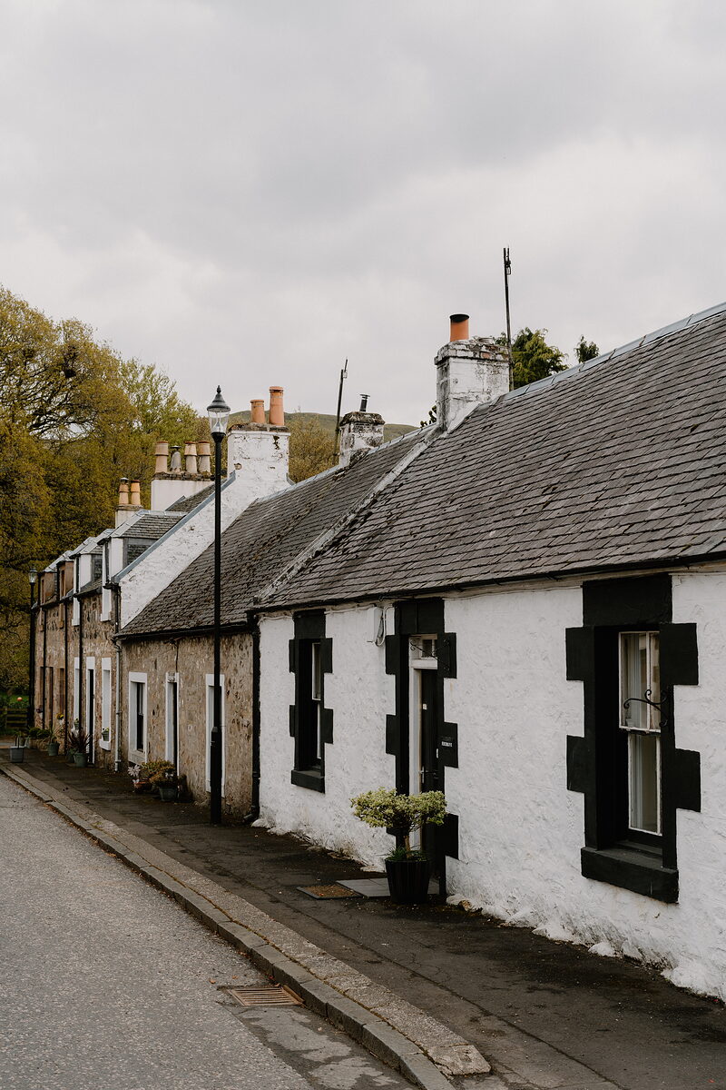 A row of whitewashed cottages with dark window surrounds lines a street, a lamppost standing nearby and a tree-covered hill rising behind.