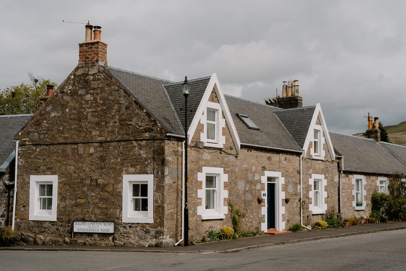 A row of stone cottages with pointed gables and white door surrounds lines a street, a street sign fixed to the corner wall.