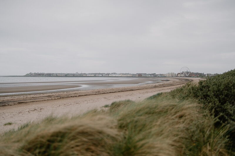 A long sandy beach stretches towards a distant seafront town, a ferris wheel rising above the rooftops and dune grass in the foreground.