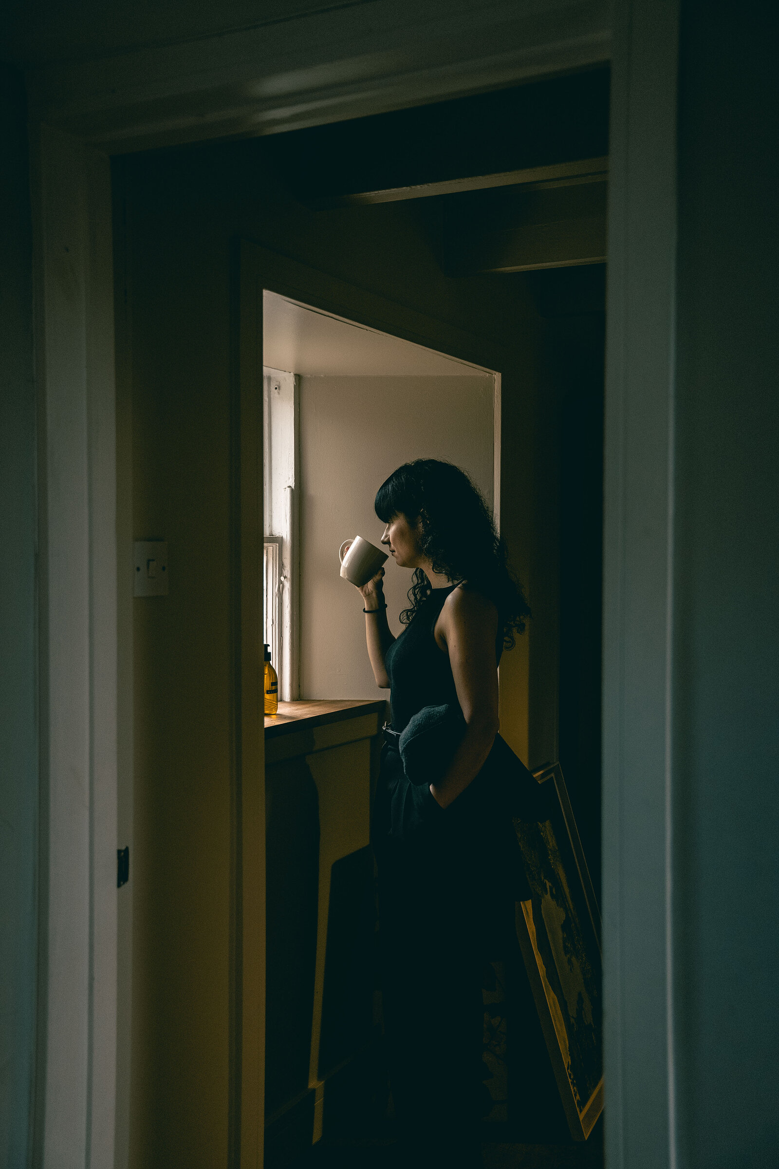 A woman with dark curly hair stands at a small window in a dim room, drinking from a mug, seen through a doorway.