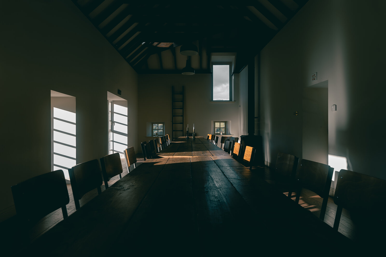 A dark dining hall at Ballintaggart with a long wooden table, rows of chairs, whitewashed walls and sunlight falling through small windows.
