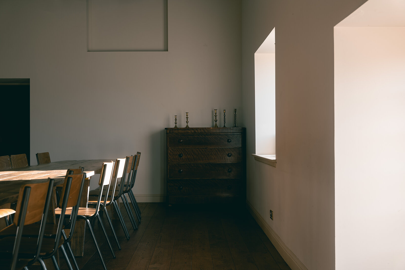 A dining room at Ballintaggart with a long wooden table, school-style chairs, a dark chest of drawers and candlesticks beside a bright window.