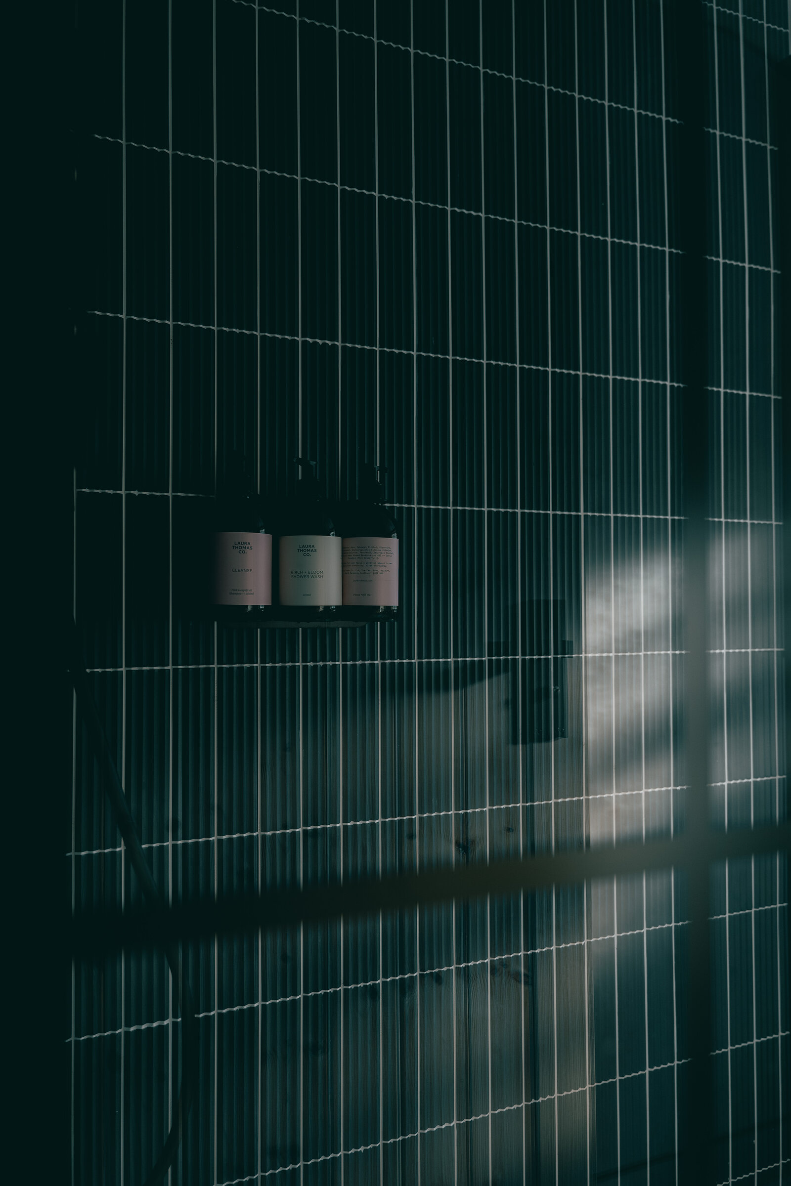 Three toiletry bottles on a small shelf against a dark green tiled shower wall, with soft light and shadow falling across it.