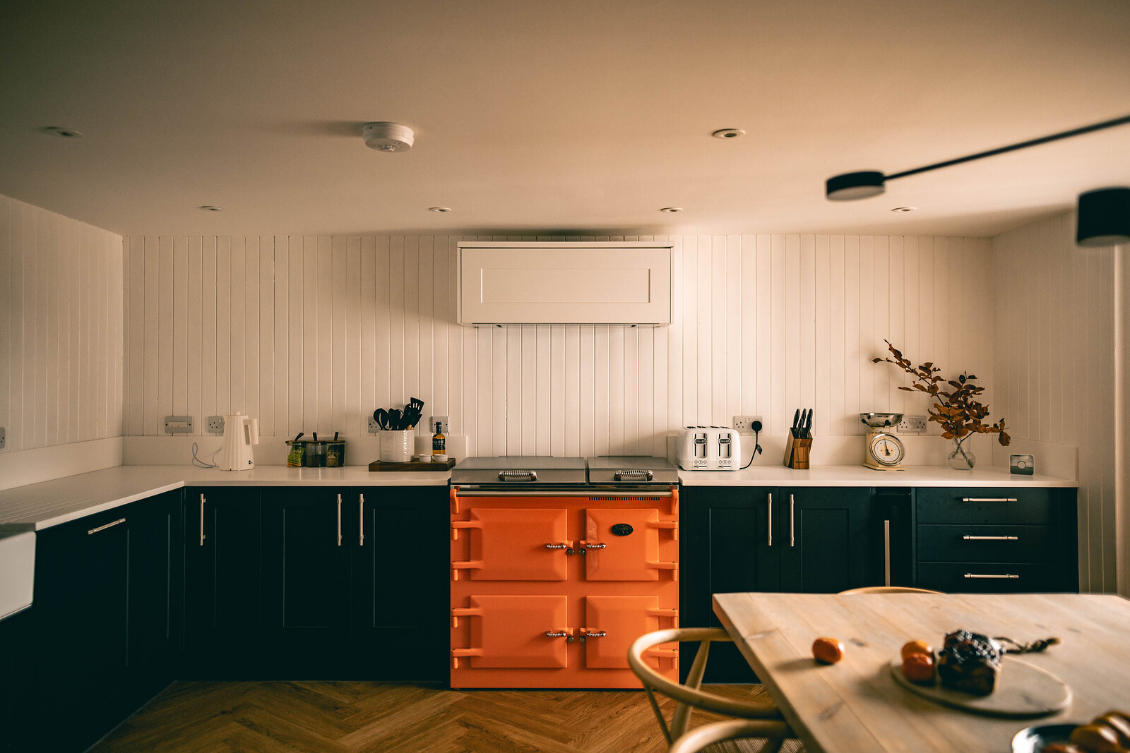 A kitchen with an orange range cooker set between dark navy cupboards, white panelled walls, a wooden table and herringbone floor.