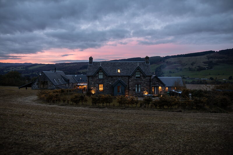 Ballintaggart farmhouse at dusk with lit windows, under a pink and grey sky, with wooded hills and fields behind.