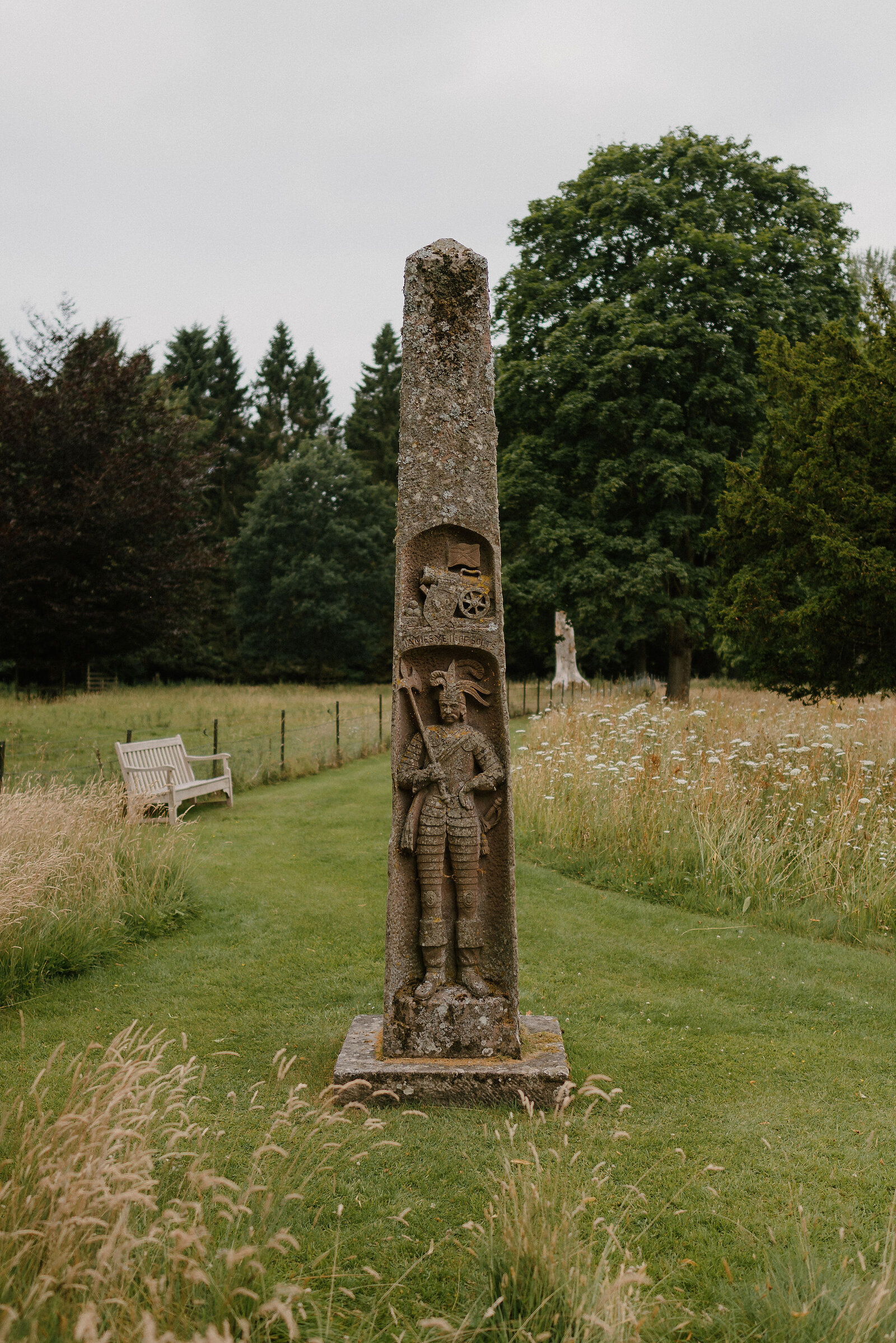 A tall carved stone pillar bearing a heraldic shield and a robed figure holding a staff, standing in a mown lawn beside a wooden bench, with tall grass, wildflowers and trees behind.