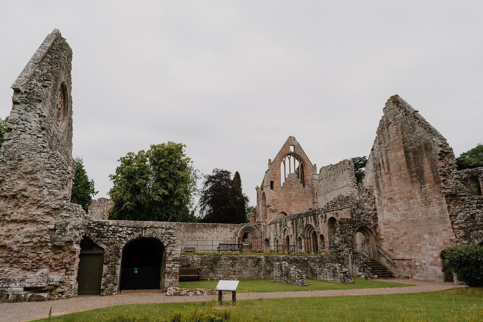 The ruined nave and transept of an abbey rising in pink sandstone above a mown lawn, with a low interpretation panel in the foreground and mature trees behind the surviving walls.
