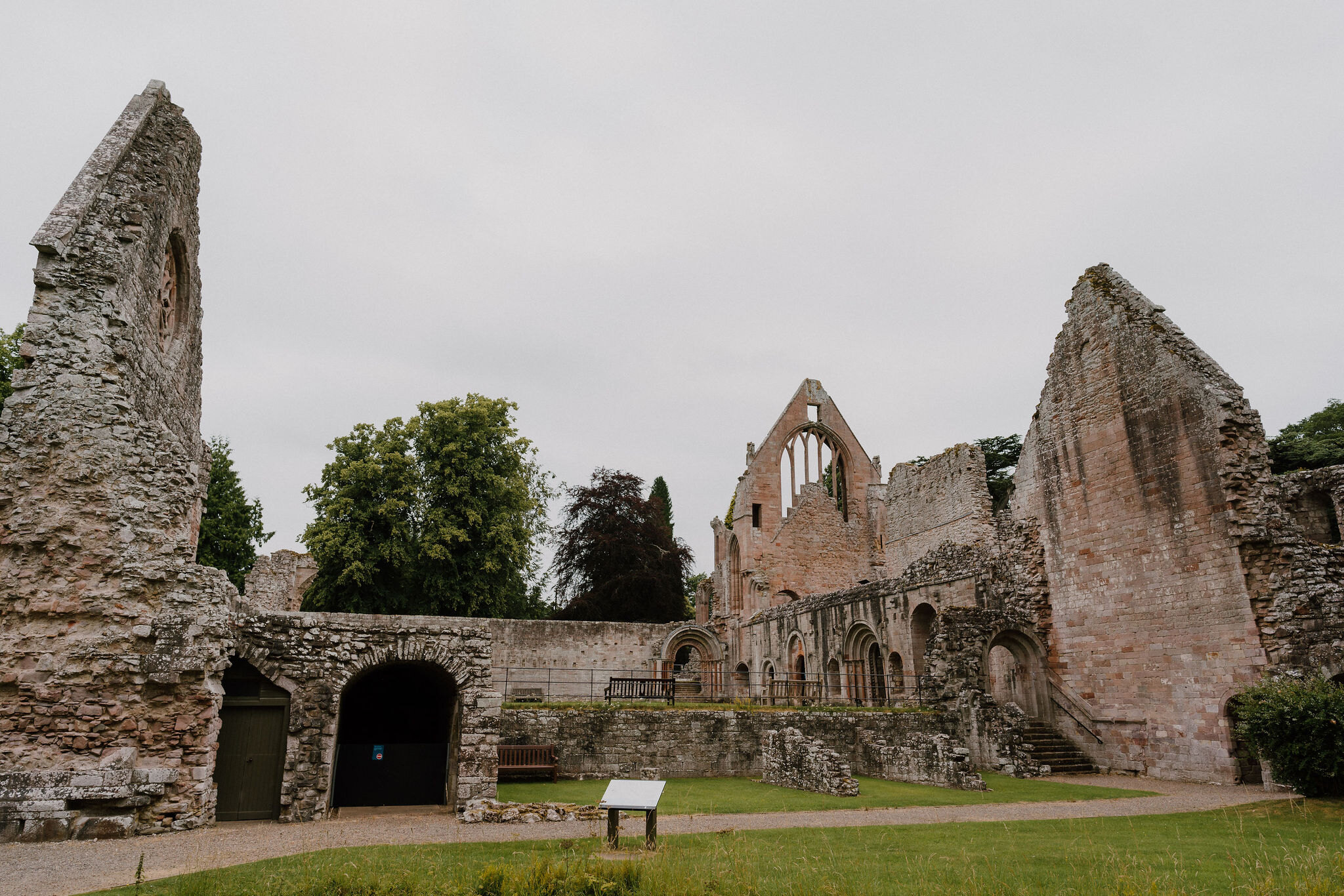 The ruined nave and transept of an abbey rising in pink sandstone above a mown lawn, with a low interpretation panel in the foreground and mature trees behind the surviving walls.