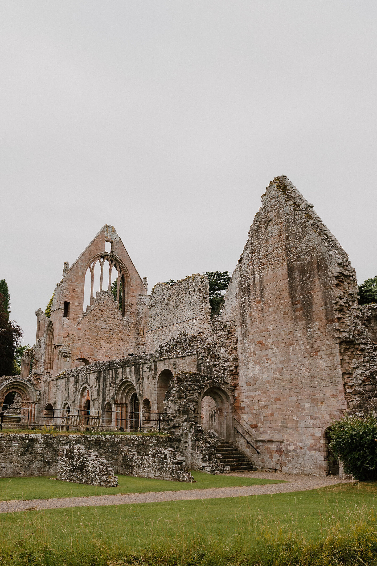 The ruined abbey church stands in weathered sandstone, a tall gothic gable and window at its centre, round-arched arcades and a stone stair leading up through the walls, lawn spreading in front.