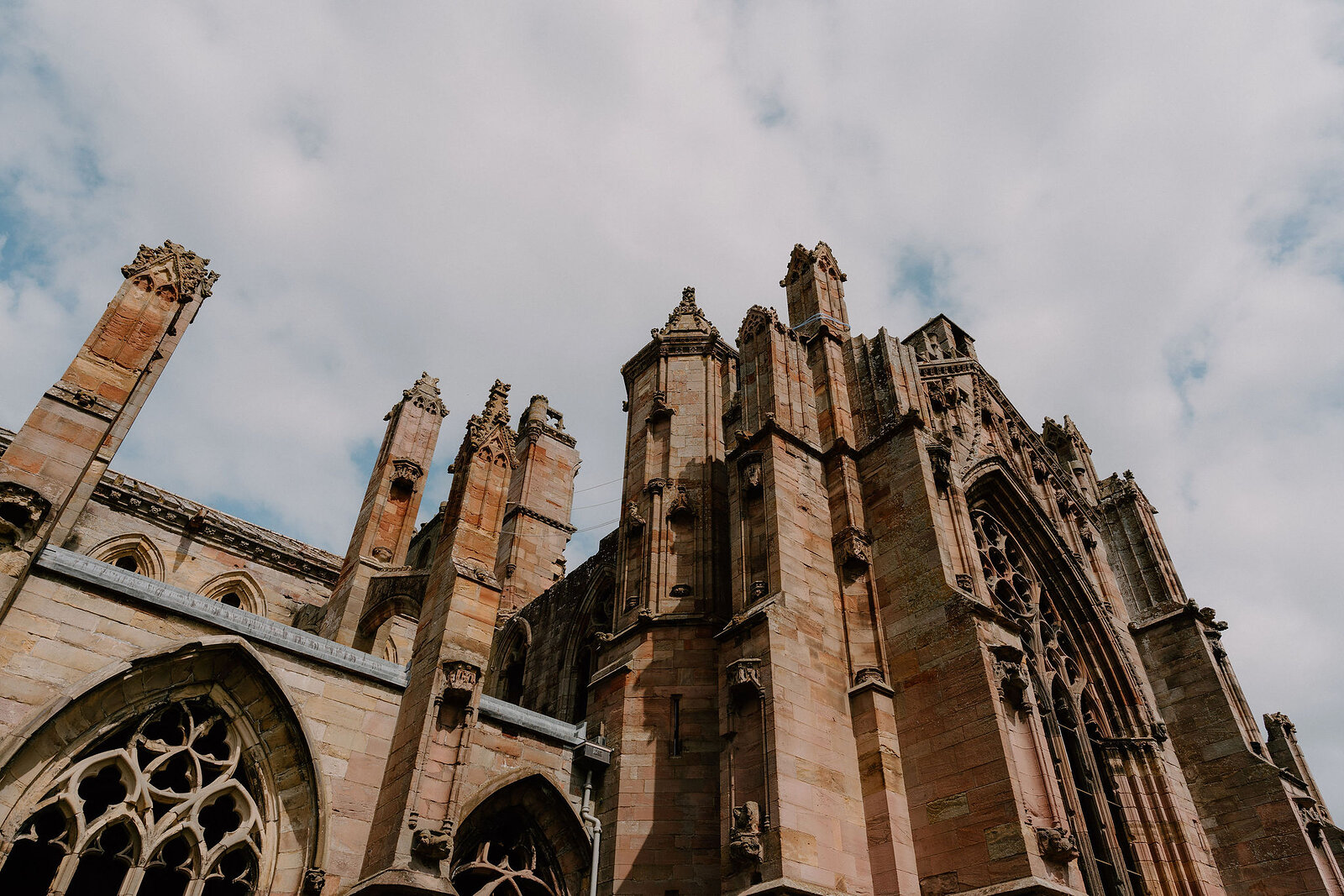 A ruined abbey's pinnacled west front rises against a cloudy sky, fragments of a gothic doorway and rose window visible below.