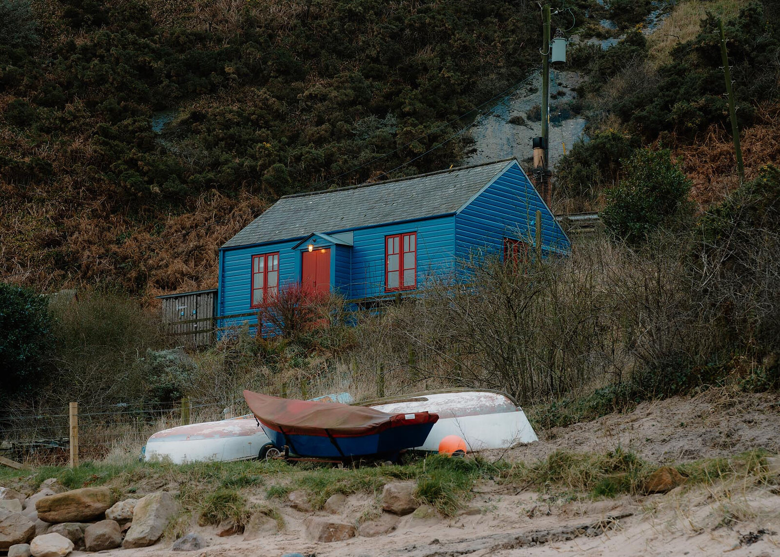 A landscape view of the Blue Cabin at the foot of the brae, lamps lit by its red door and boats drawn up on the grass.