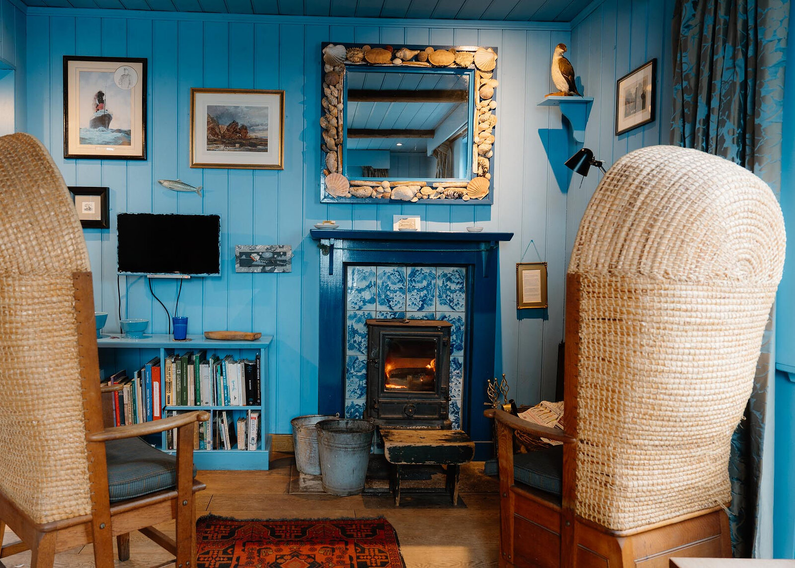 A landscape view of the living room: two straw-hooded Orkney chairs framing the stove, bookshelves, pictures and a shell mirror on the blue boards.