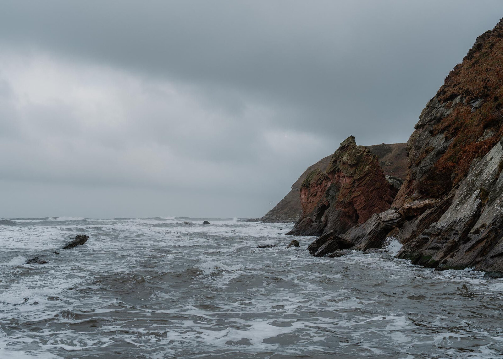 Waves swirl round the dark rocks and sea stacks at the mouth of the cove under a heavy sky.