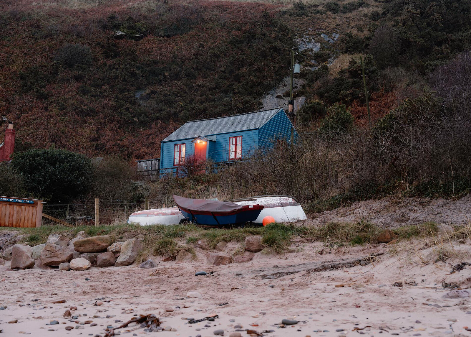 A landscape view of the Blue Cabin at dusk from the beach, its door lamps lit and boats on the grass below.
