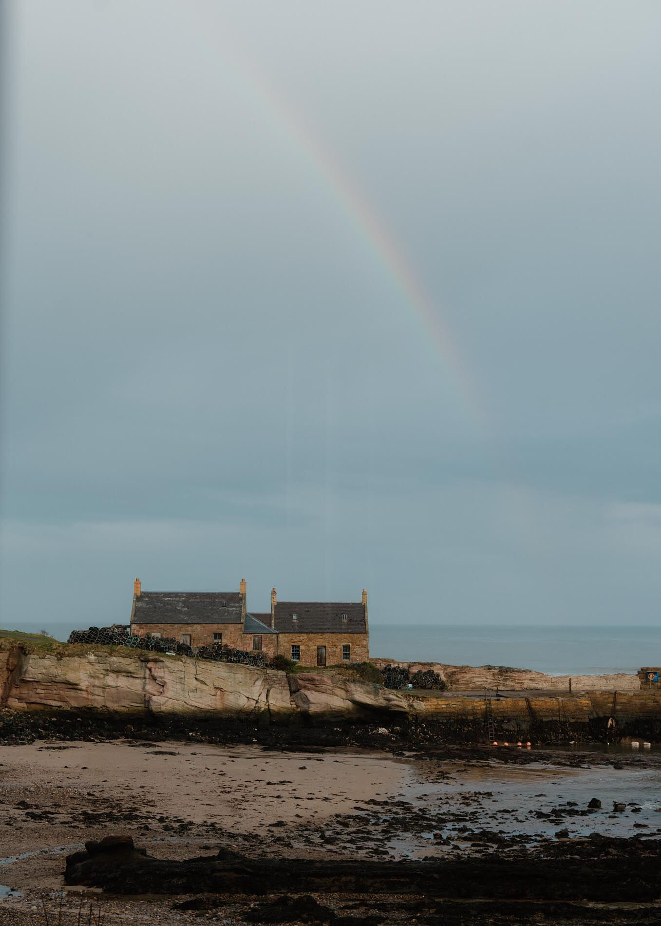 A rainbow arcs down behind the stone houses on the pier, the rocks and sand of the harbour at low water.