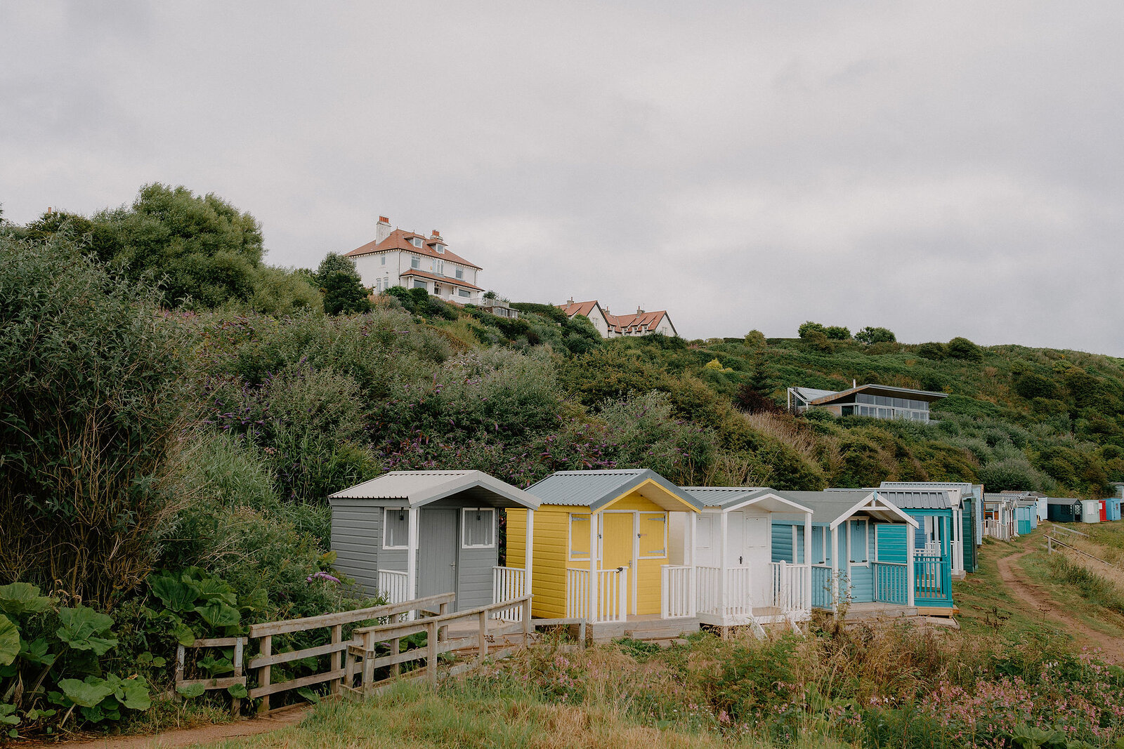 A row of colourful beach huts lines a grassy slope, a large house visible on the hilltop above.