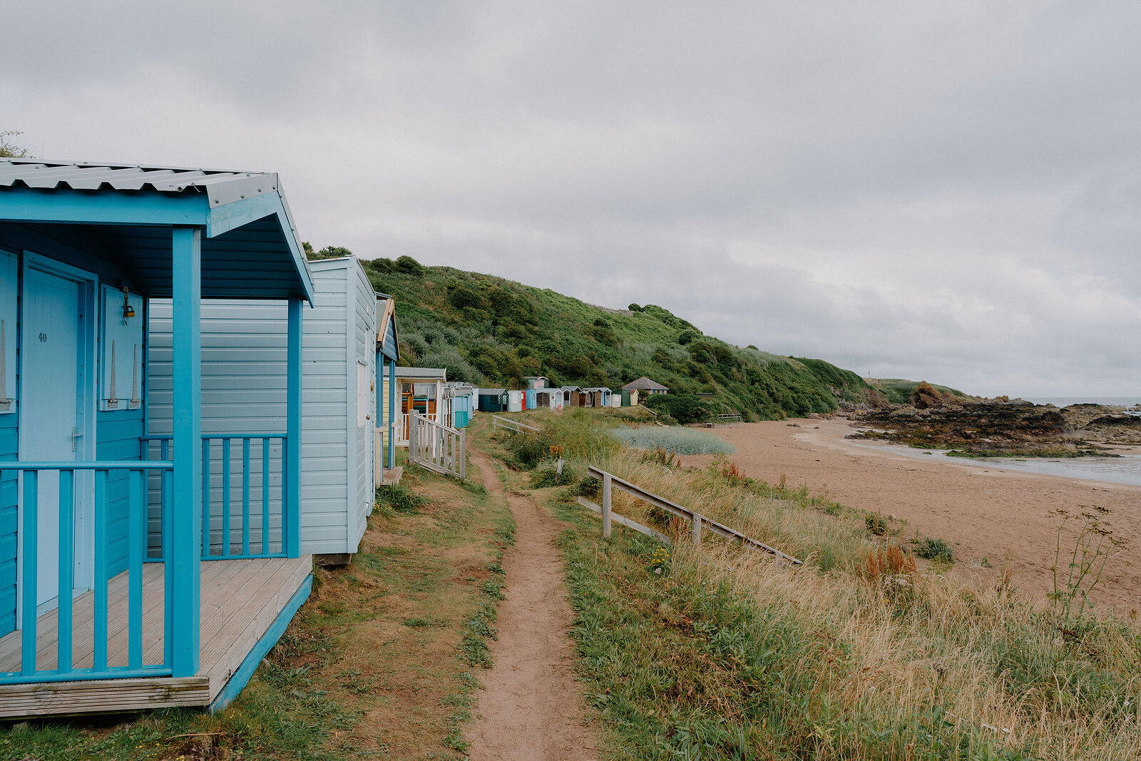 A row of pastel-coloured beach huts lines a path above a sandy beach, a headland and open sea visible beyond.