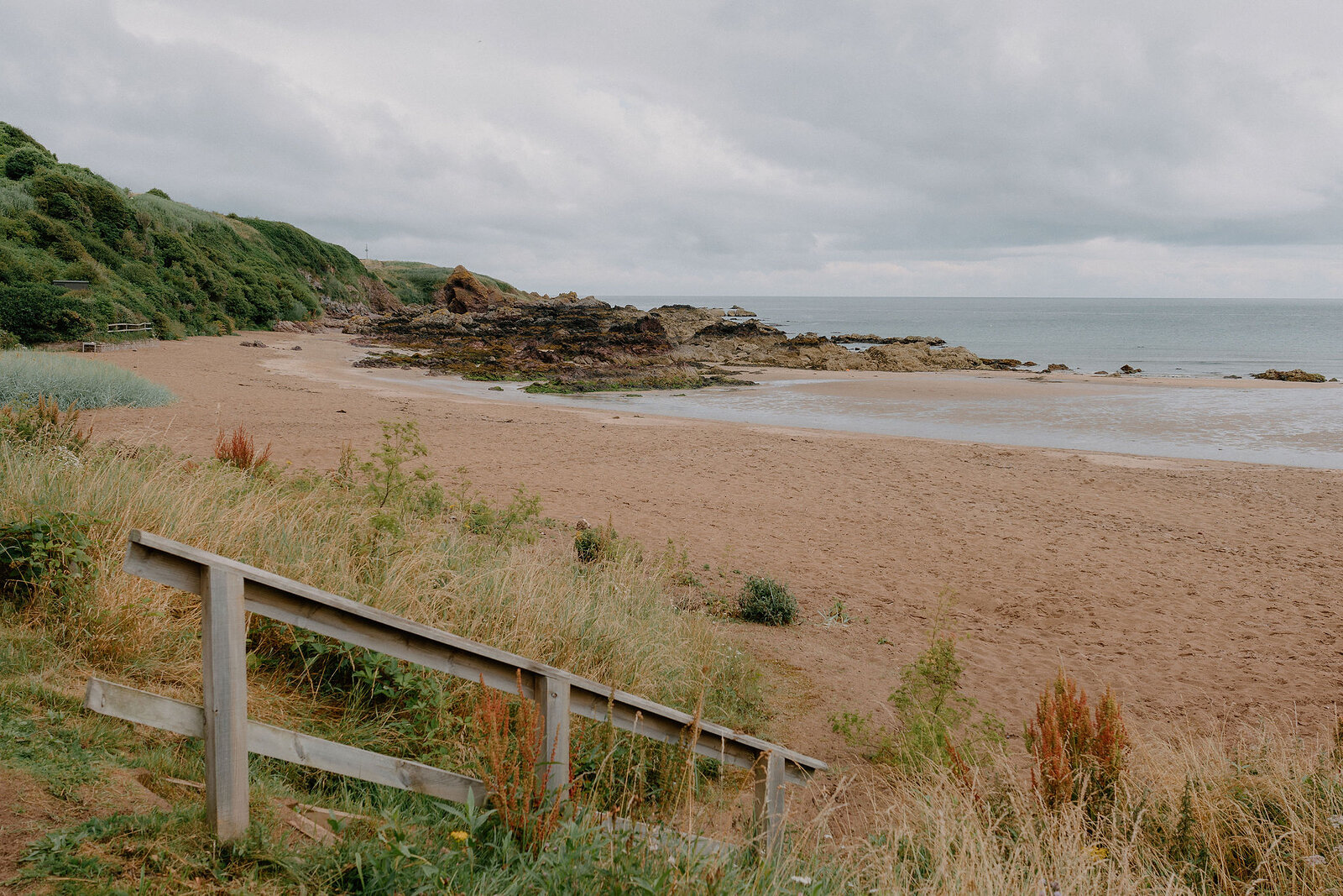 A wooden fence and flowering gorse line the top of a sandy beach, a rocky headland with a small hut visible and open sea beyond.