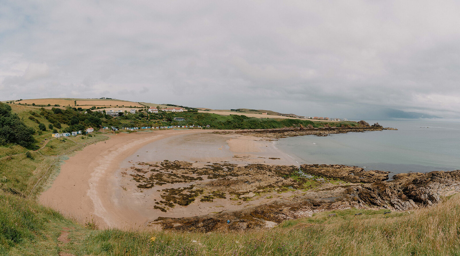 A panoramic view shows a sandy cove backed by low hills, houses scattered along the shore and rocks breaking the water in the foreground.