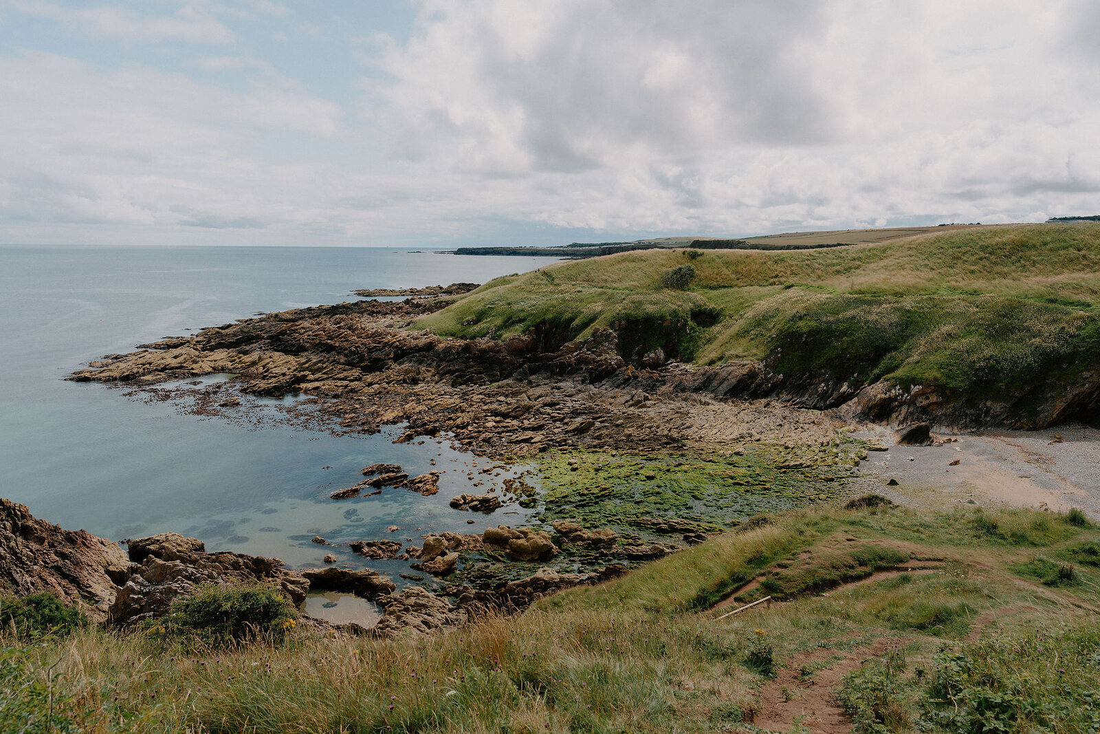 A rocky cove edged with seaweed and grassy headland lies beside calm open water, a sandy strip curving along the shore.