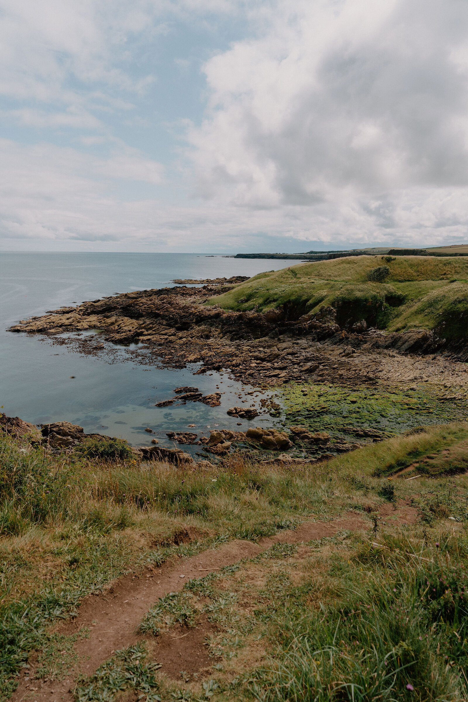 A rocky cove streaked with green seaweed curves beside calm water, a grassy headland and coastal path leading down to the shore.