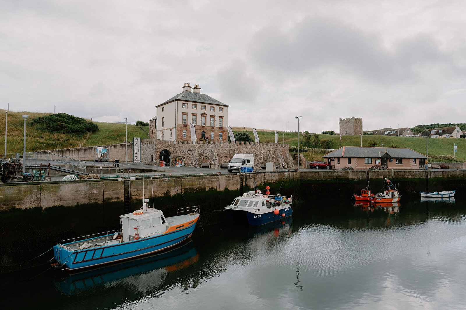 A grand pavilion building with tall chimneys overlooks a small harbour, fishing boats and gulls on the still water in front.
